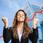 excited business woman in front of skyscrapers with sales arrows behind her trending up